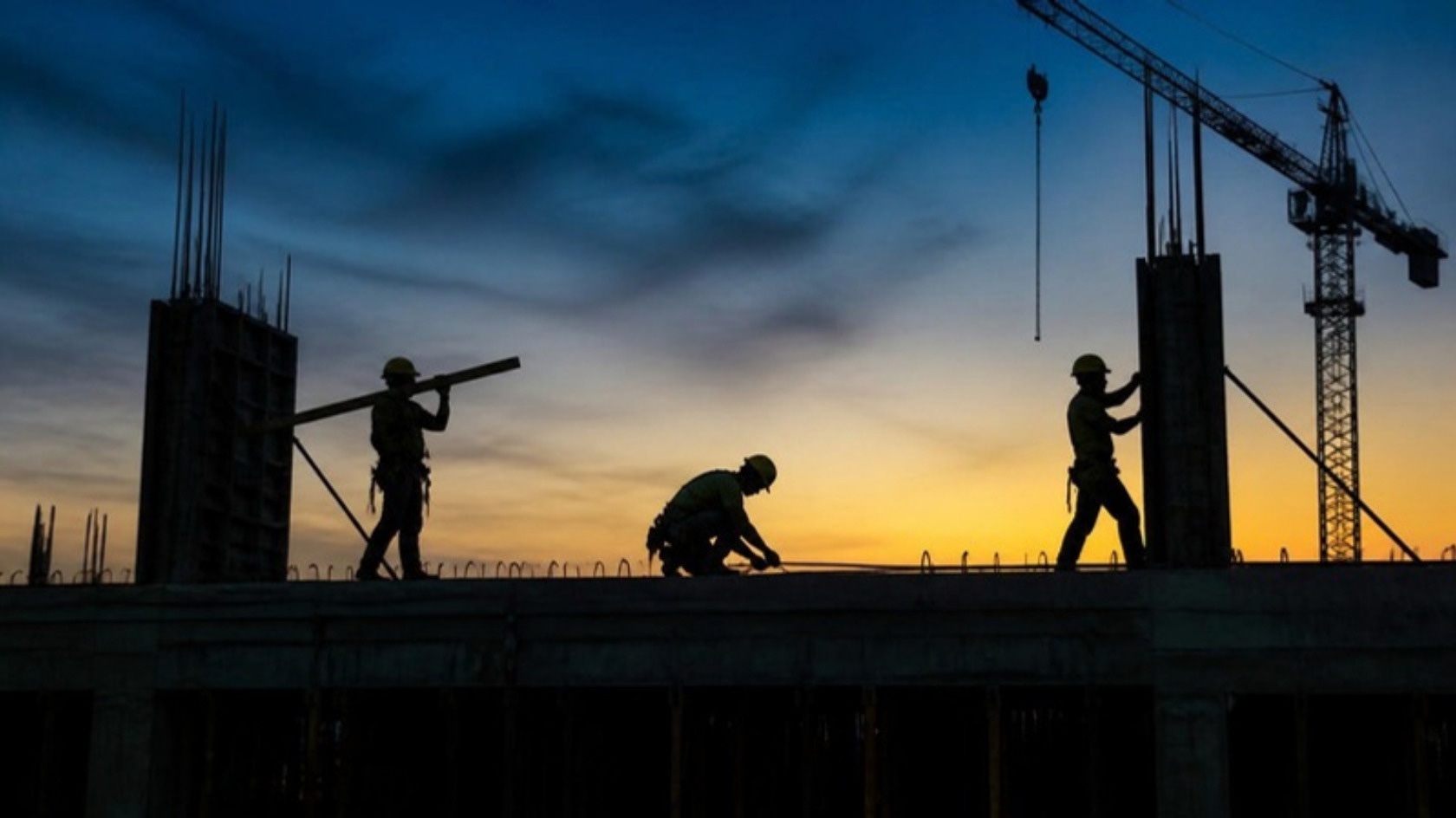 Forming crew working on a concrete deck at dusk, with a tower crane behind